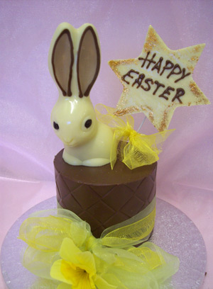 a picture of one white chocolate bunny rabbit on a milk chocolate tier. Decorated with yellow flowers and mounted on a silver display disc.