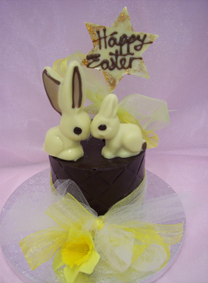 a picture of two white chocolate bunny rabbits on a milk chocolate tier. Decorated with yellow flowers and mounted on a silver display disc.