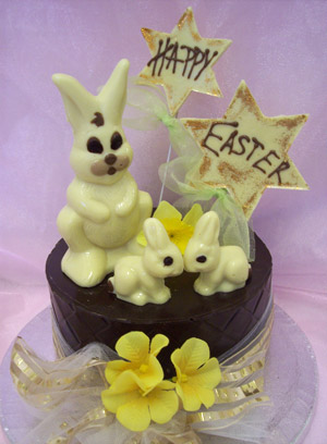 a picture of three white chocolate bunny rabbits on a milk chocolate tier.  Decorated with yellow flowers and mounted on a silver display disc.