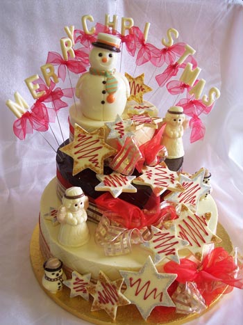 a picture of a white chocolate snowman with Angels decorated with coloured chocolate, snowflakes and red ribbon on chocolate tiers.  Chocolate letters reads 'Merry Christmas'
